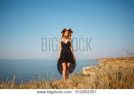 The Girl In A Black Dress And Hat On A Rocky Beach Looks At The Sea