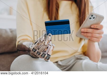 Woman With Contemporary Prosthesis Arm Holds Credit Card And Mobile Phone Sitting On Sofa In Living 