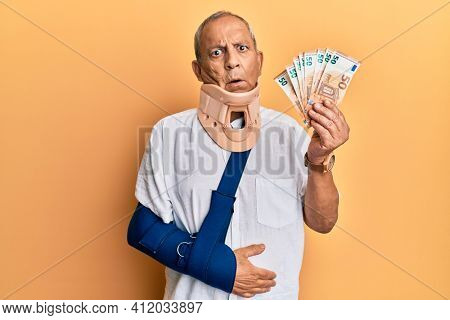Handsome mature injured man wearing cervical collar and sling holding insurance euros in shock face, looking skeptical and sarcastic, surprised with open mouth 