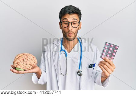 Young handsome man holding brain and pills as mental health concept making fish face with mouth and squinting eyes, crazy and comical. 
