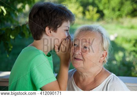 Boy whispering to grandma in summer outdoors garden, grandkid and grandmother lalking.