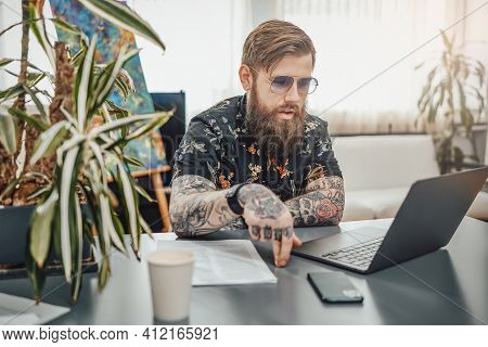 Cosy Home Office And Remote Working. Man With Beard And Tattooed Body Wearing Stylish Clothing Doing