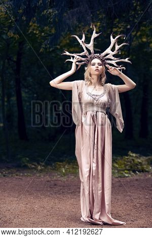 Sensual Caucasian Girl Posing With Artistic Deer Horns And Long Light Dress In Summer Forest