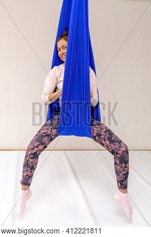 A Young Woman Poses While Doing Anti-gravity Aerial Yoga In A Blue Hammock On A White Background.