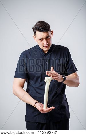Young Male Doctor Chief Physician In A Black Surgical Suit. Close-up Portrait. Holds A Femur In His
