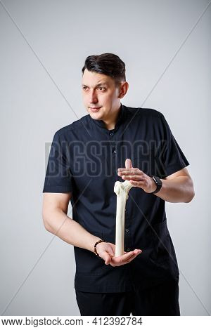 Young Male Doctor Chief Physician In A Black Surgical Suit. Close-up Portrait. Holds A Femur In His
