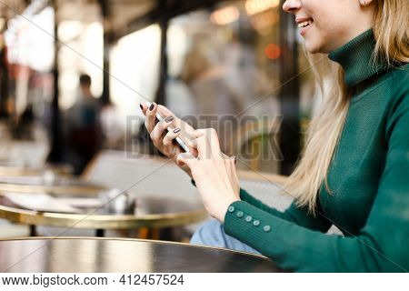 Blonde Girl Chatting By Smartphne And Sitting At Street Cafe.