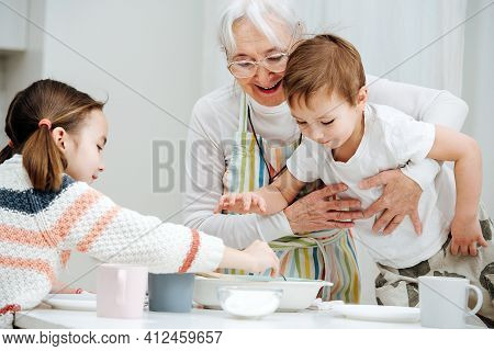 Happy Granny Holding Her Grandson From Falling. He Is Standing On A Stool, Reaching For The Food. Hi