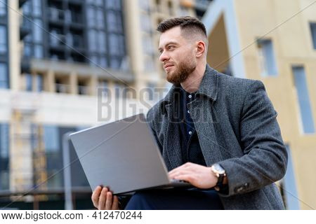 Portrait Of A Handsome Young Man Working On Laptop Outdoors. Fashion Man Sits On Bench In City Cente