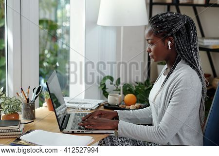 Serious Young Black Woman With Dreadlocks Working On Laptop At Her Desk At Home And Writing Essay Fo