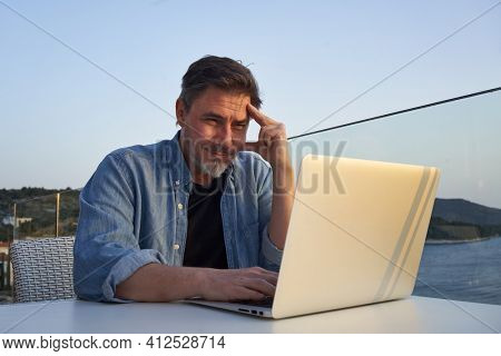 Happy man working online with laptop computer on terrace. Home office, browsing internet, tele working, video chat. Businessman on vacation at sea. Escape, freedom, getaway.