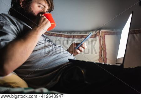 Bearded Man Consulting Her Phone While Enjoys Watching His Favorite Series On His Laptop At Night. P