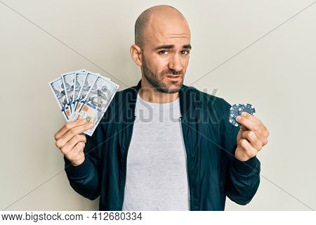 Young hispanic man playing poker holding dollars banknotes and chips clueless and confused expression. doubt concept. 