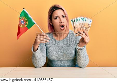Hispanic woman with pink hair holding portugal flag and euros banknotes in shock face, looking skeptical and sarcastic, surprised with open mouth 