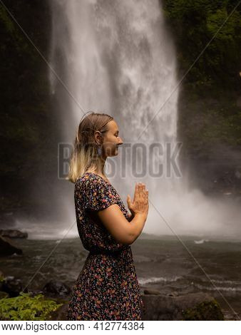 Young Caucasian Woman Spending Time Near Waterfall. Meditation And Relaxation. Hands In Namaste Mudr