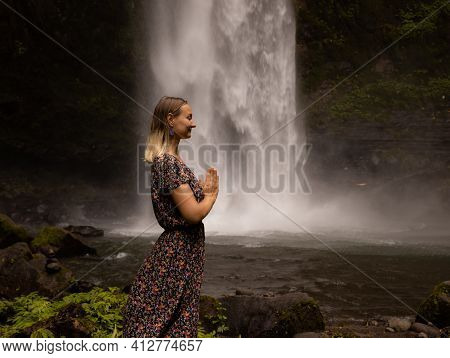 Young Caucasian Woman Spending Time Near Waterfall. Meditation And Relaxation. Hands In Namaste Mudr