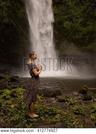 Young Caucasian Woman Spending Time Near Waterfall. Meditation And Relaxation. Hands In Namaste Mudr