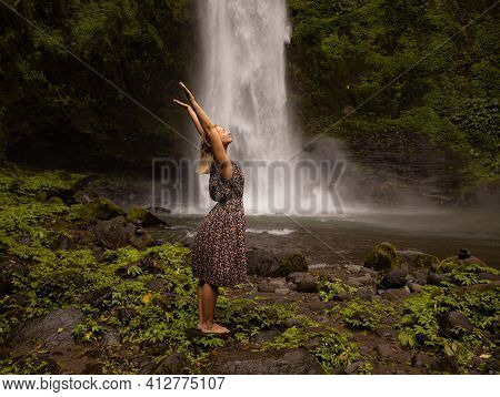 Happy Caucasian Woman Spending Time Near Waterfall, Raising Hands Up. Nature Concept. Energy Of Wate