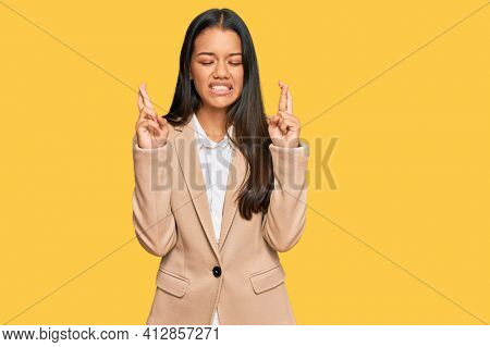 Beautiful hispanic woman wearing business jacket gesturing finger crossed smiling with hope and eyes closed. luck and superstitious concept. 