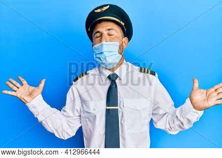 Handsome man with beard wearing airplane pilot uniform wearing safety mask celebrating mad and crazy for success with arms raised and closed eyes screaming excited. winner concept 