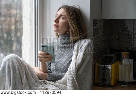 Pensive Woman Wrapped In Knitted Plaid Sitting On Windowsill Looking Through The Window At Home, Thi