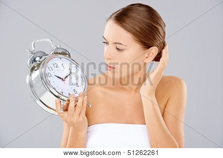 Beautiful young woman looking at a large silver retro alarm clock with bells that she is holding in consternation as though waiting for it to ring