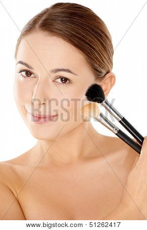 Beautiful naked young woman with neatly tied back brunette hair and a gentle smile holding two large soft cosmetic brushes up against her cheek