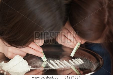 Two girls snorting an illegal white powder with dollar bills off a mirror