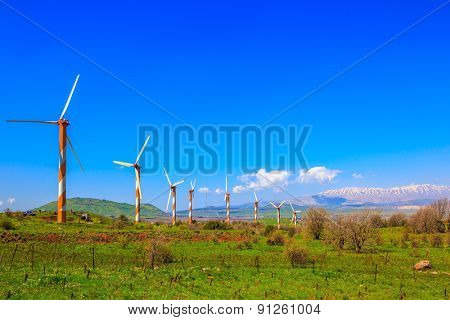  Several huge modern windmills on background snowy Hermon. Flowering Golan Heights on a sunny day. Galilee, Israel