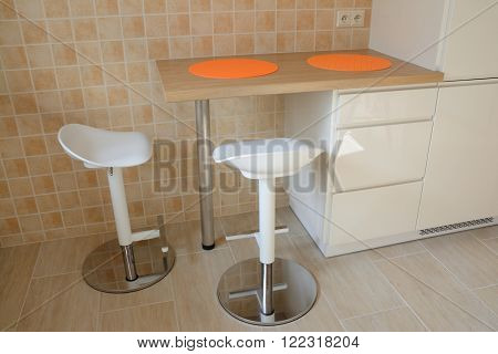 Two swivel stools and kitchen small table with orange rubber tablemats at wall covered with tiles