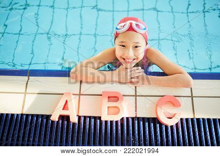 Happy kid in swimwear looking at camera with smile while standing in water by edge of swimming-pool with light letters A, B and C near by