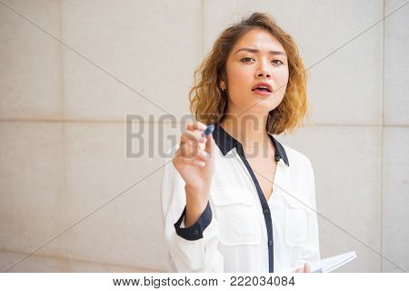 Portrait of exited young Asian woman with notebook pointing pen at camera. Writer finding ideas for new article. Writing and inspiration concept
