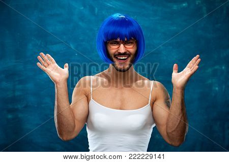 Happy smiling young man wearing blue wig and female cloth. Cross dressing male person posing on blue background.