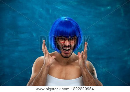 Angry frustrated young man wearing blue wig and female cloth. Cross dressing male person posing on blue background.