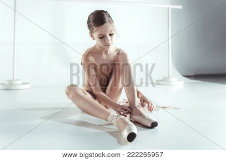 Beautiful little ballerina sitting on the floor and puting on foot pointe shoes on white ballet studio background