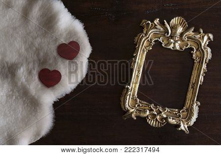 antiq golden photo frame in Rococo style, on dark rustic wooden background with white Tuscan sheepskin and hearts