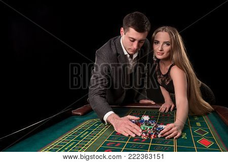Young beautiful couple takes their winnings at the roulette table at the casino, on a black background. A man in a suit with a woman in a black dress