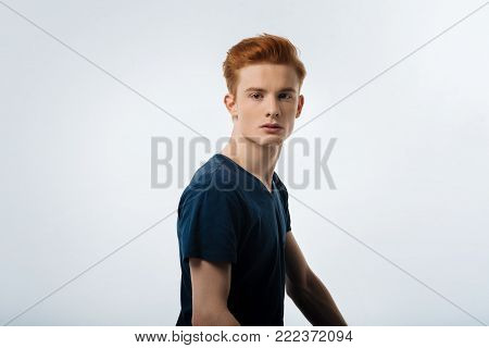 Model. Good-looking stony-faced red-haired young man having a modern hairstyle and wearing a black t-shirt and looking in the distance