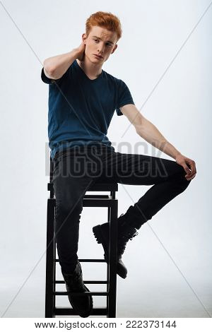 Fashionable. Handsome earnest red-headed young man sitting on the chair and thinking and having his hands on his neck and knee