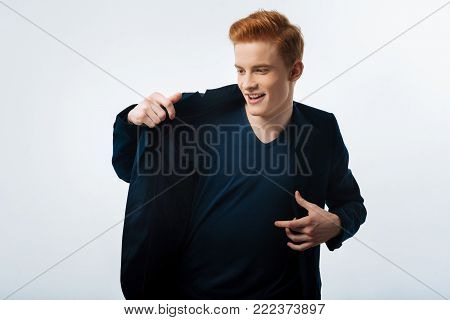 Happy. Handsome inspired red-haired young man putting on a black jacket and smiling and having a modern haircut