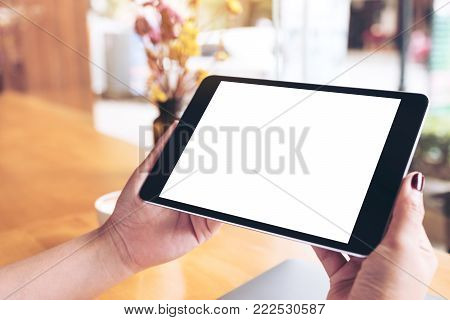 Mockup image of woman's hands holding black tablet pc with blank white desktop screen , laptop and coffee cup on wooden table in cafe