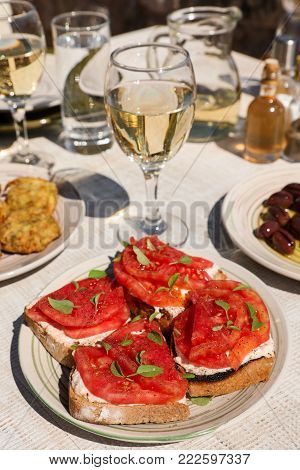Sandwiches with feta cheese, fresh tomatoes, basil leaves as an appertizer next to plates of zucchini balls, olive olives and white wine, water glasses served in greek tavern. Vertical. Close-up.