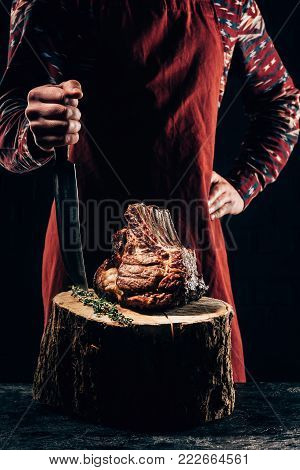 cropped shot of chef in apron standing with meat knife and delicious grilled ribs on wooden stump