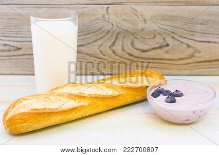 Healthy Breakfast Concept with a glass of milk, youghurt with fresh berries and a french baguette on a light wooden table against rustic wooden wall