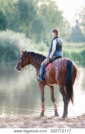 Young teenage girl equestrian riding horseback to river at early morning in rays of sunlight. View from backside. Multicolored vibrant outdoors vertical summertime image with vintage filter.