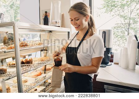 Portrait of white Caucasian beautiful barista woman taking muffin pastry from shop-window. Person at work place small business concept