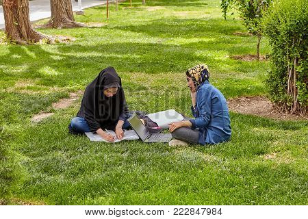 Isfahan, Iran - April 24, 2017: Two Muslim female students, dressed in Islamic hijab, are preparing for the exams, sitting on the lawn in the city park, using a laptop.
