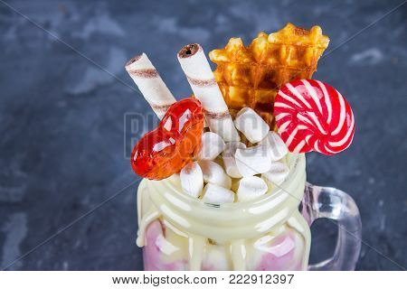 Freakshake from pink smoothie, cream. Monstershake with lollipops, waffles and marshmallow. Extreme milkshake in a Mason jar. Gray dark concrete background