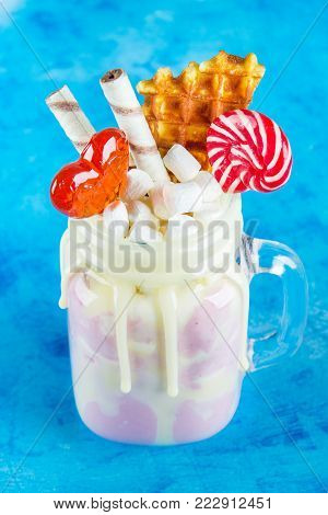 Freakshake from pink smoothie, cream. Monstershake with lollipops, waffles and marshmallow. Extreme milkshake in a Mason jar. Blue concrete background