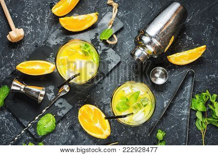 Cold alcoholic summer citrus cocktail with orange and mint in glasses and on dark stone background. Cocktail making bar tools, shaker, glasses, mint leaves. Top view